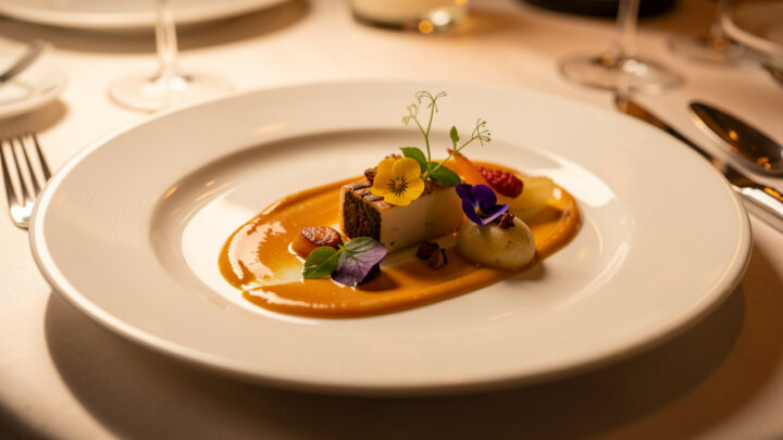 Assiette de haute gastronomie française au dressage minimaliste, accompagnée d’une sauce brillante et de fleurs comestibles sous une lumière chaude.